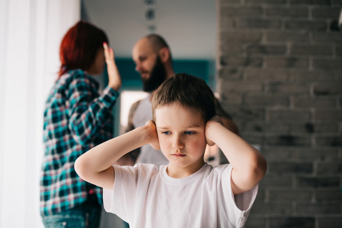 child with hands over ears with parents in background