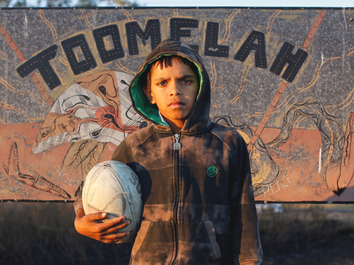 Still from Toomelah showing a boy holding a football in front of a side that reads 'Toomelah'