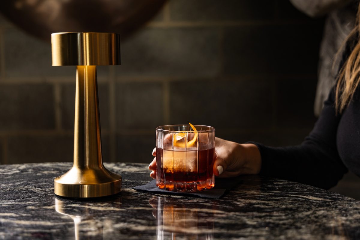A hand holds a negroni next to a brass lamp.