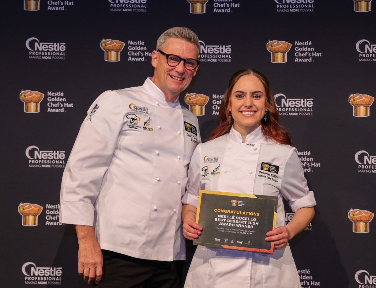 An older man in glasses and young woman holding a certificate both wear chef uniforms and smile in front of branded backdrop.