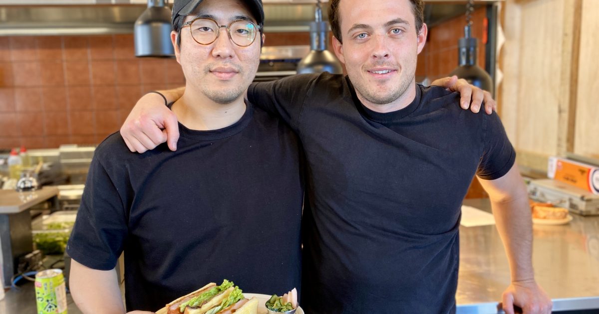 Two men in black stand in front of a kitchen holding a sandwich on a plate.