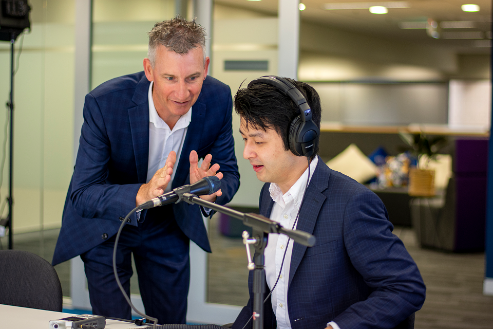 Cam Sullings talking to a person sitting at a table wearing headphones and speaking into a microphone