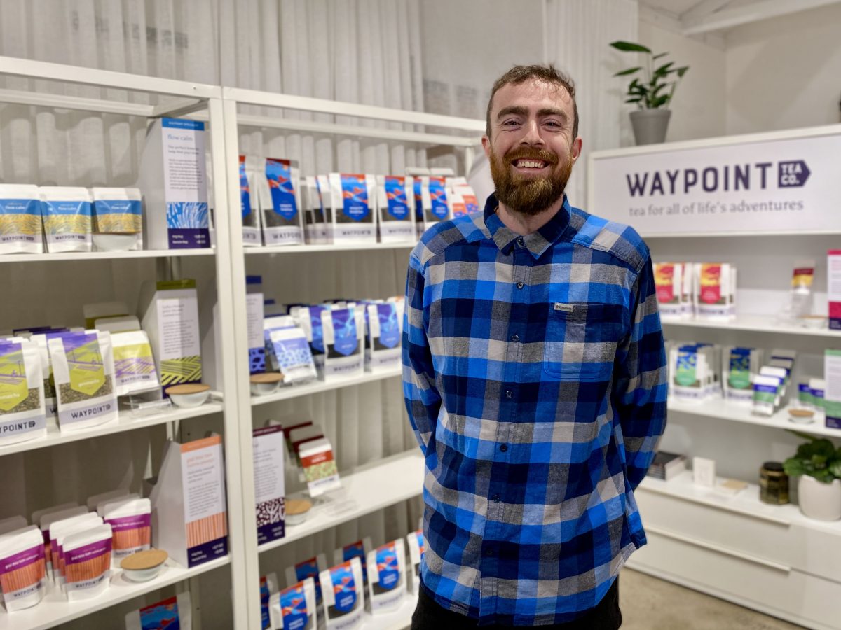 A bearded man in a blue checked shirt stands in front of shelves covered in colourful packets of tea.