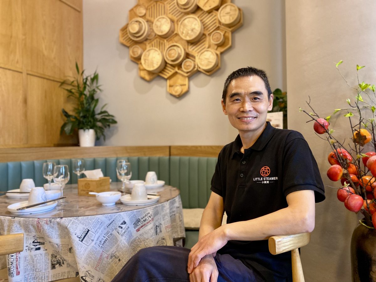 A Chinese man sits at a table set with plates and bowls, on the wall is a design of bamboo steamer baskets.