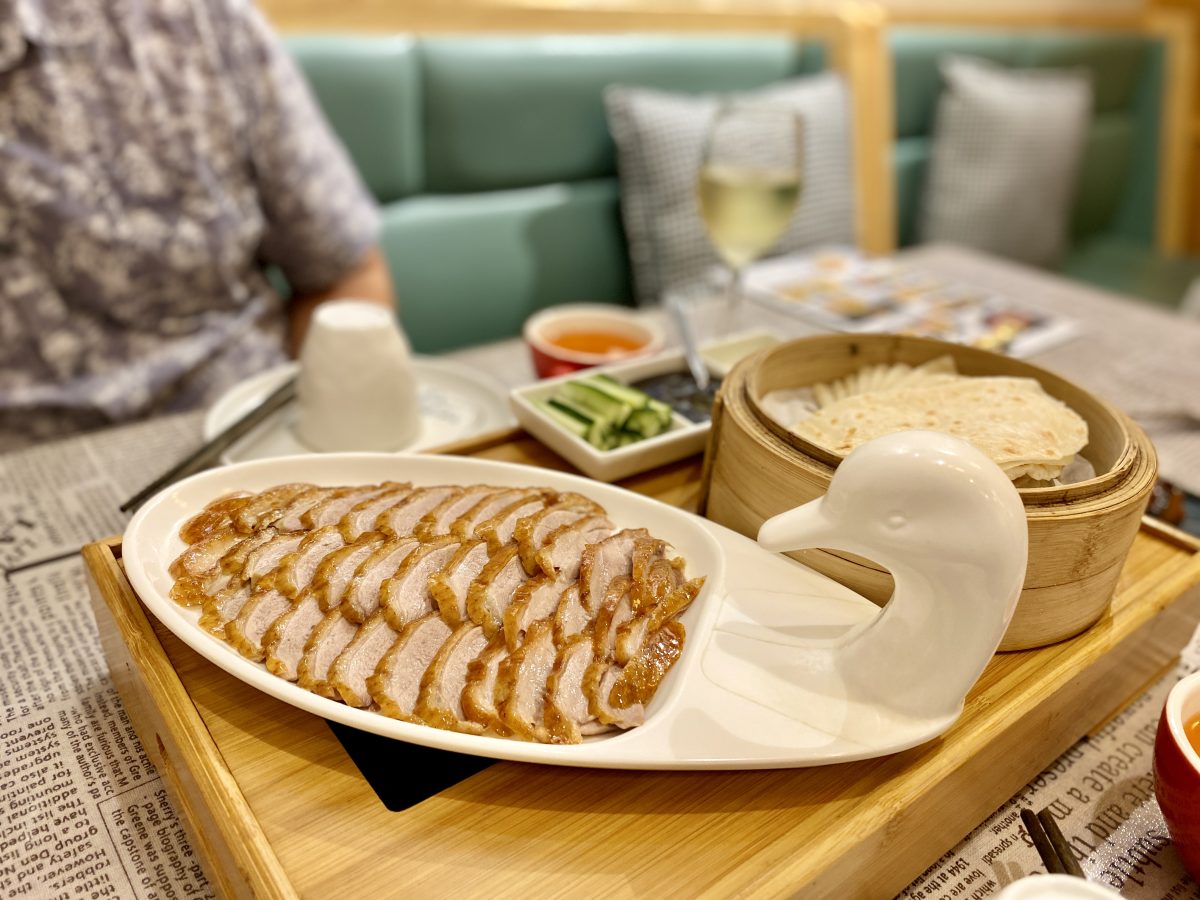 A cute white plate with a duck's head design filled with perfect slices of roast duck. Behind, a steamer basket of pancakes and other toppings.
