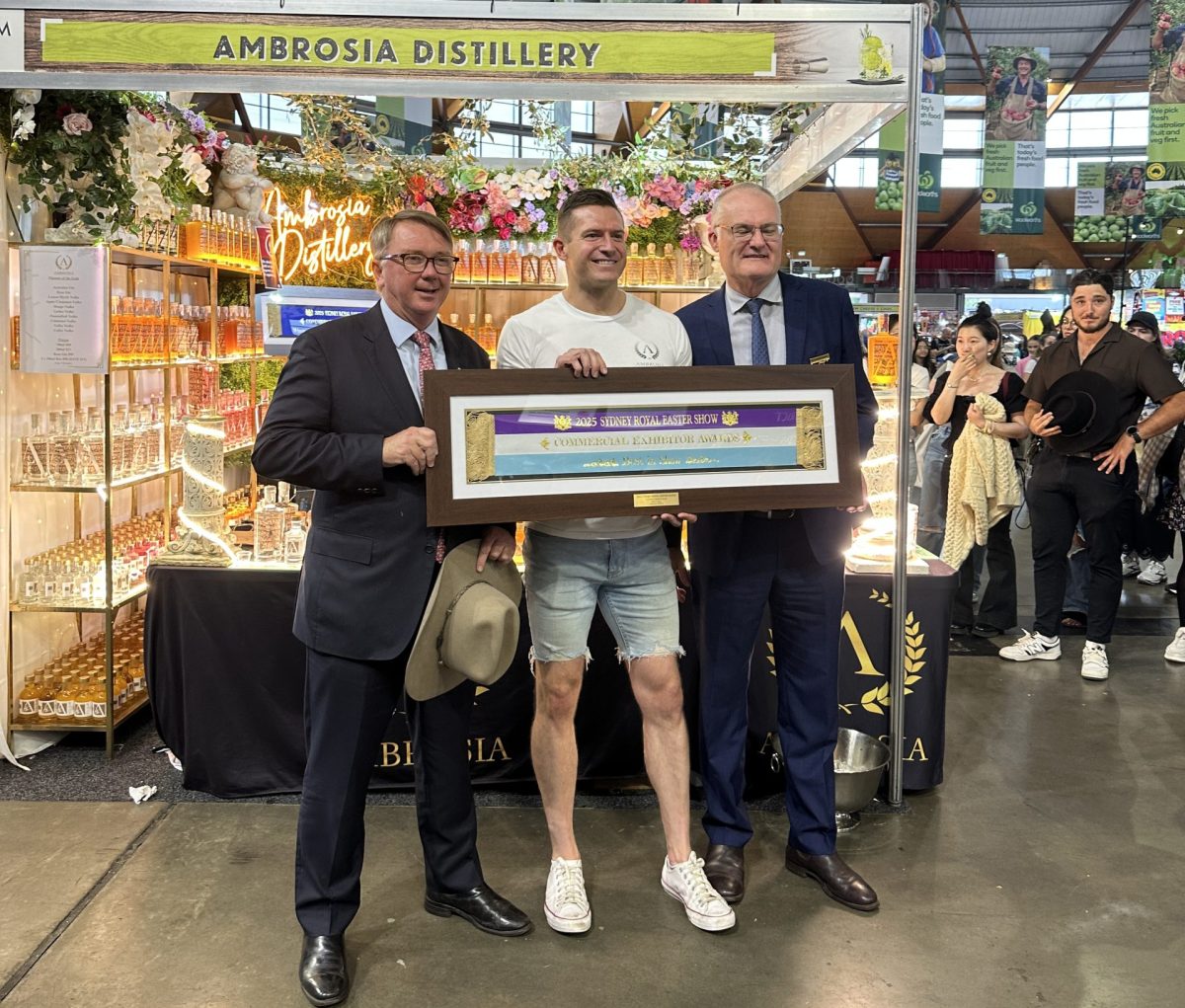 A man in shorts holds a framed ribbon award between two men in suits.