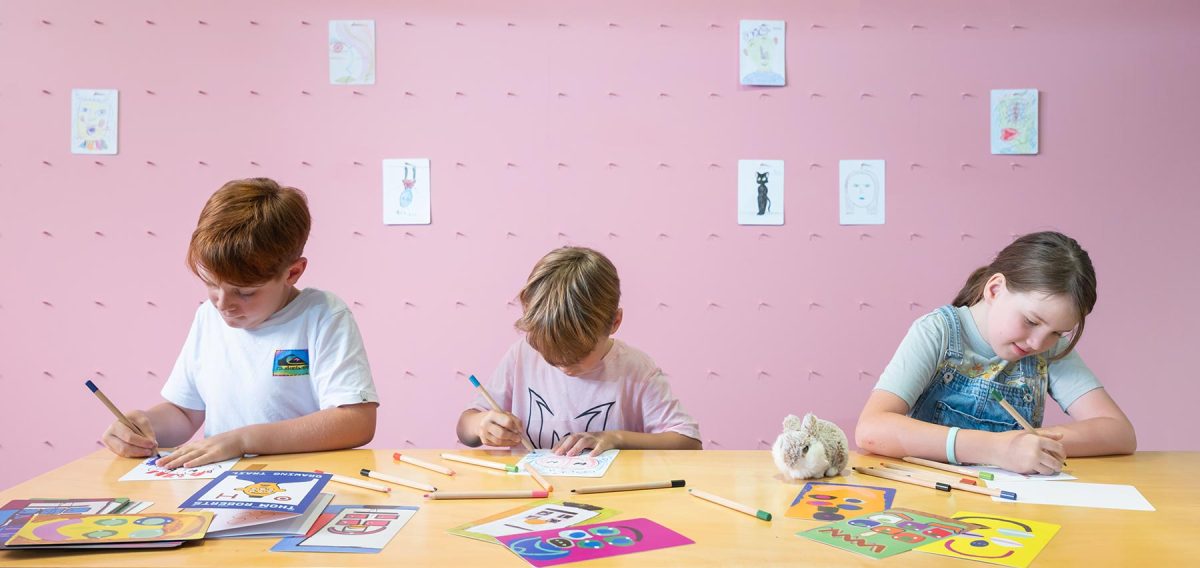 3 kids drawing on paper on a table in front of a pink wall