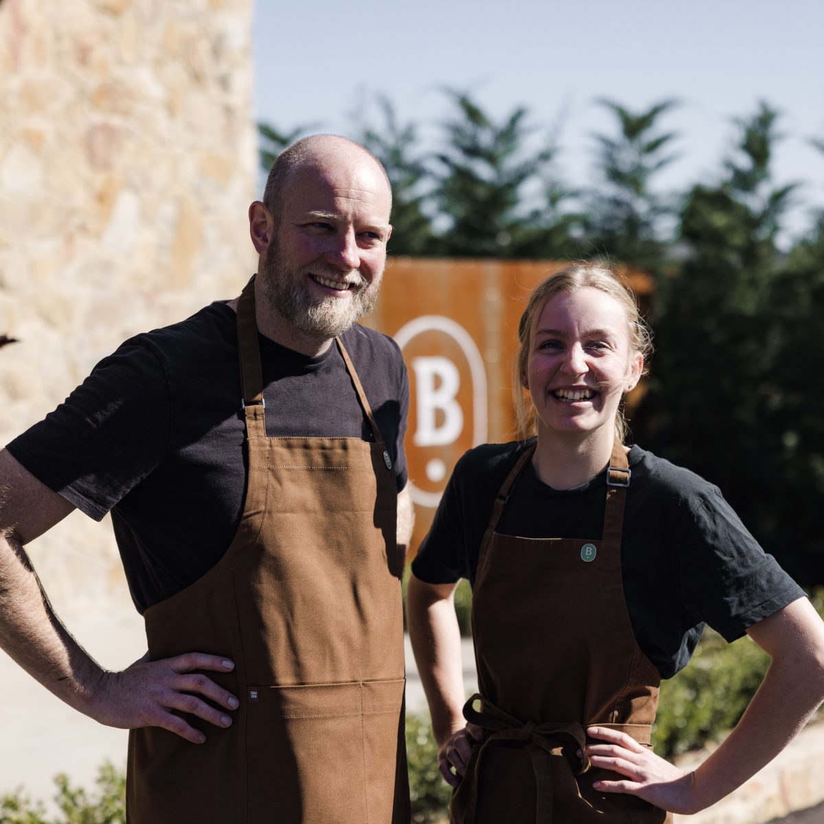 A tall man and a shorter woman - both wearing brown aprons - smile with hands on hips in front of Beltana Farm sign.