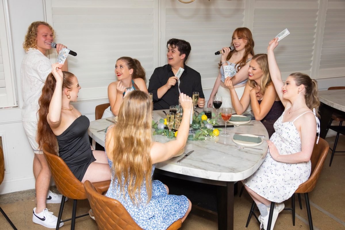 ABBA Dining Experience members at a table wave money as two hold microphones.