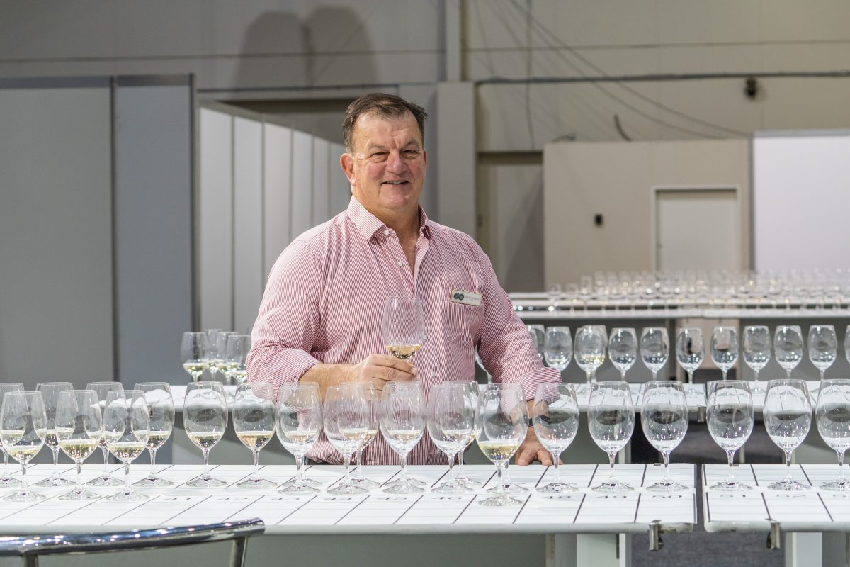 A middle-aged white man in a pink shirt stands behind a table set with many wine glasses ready for judging.