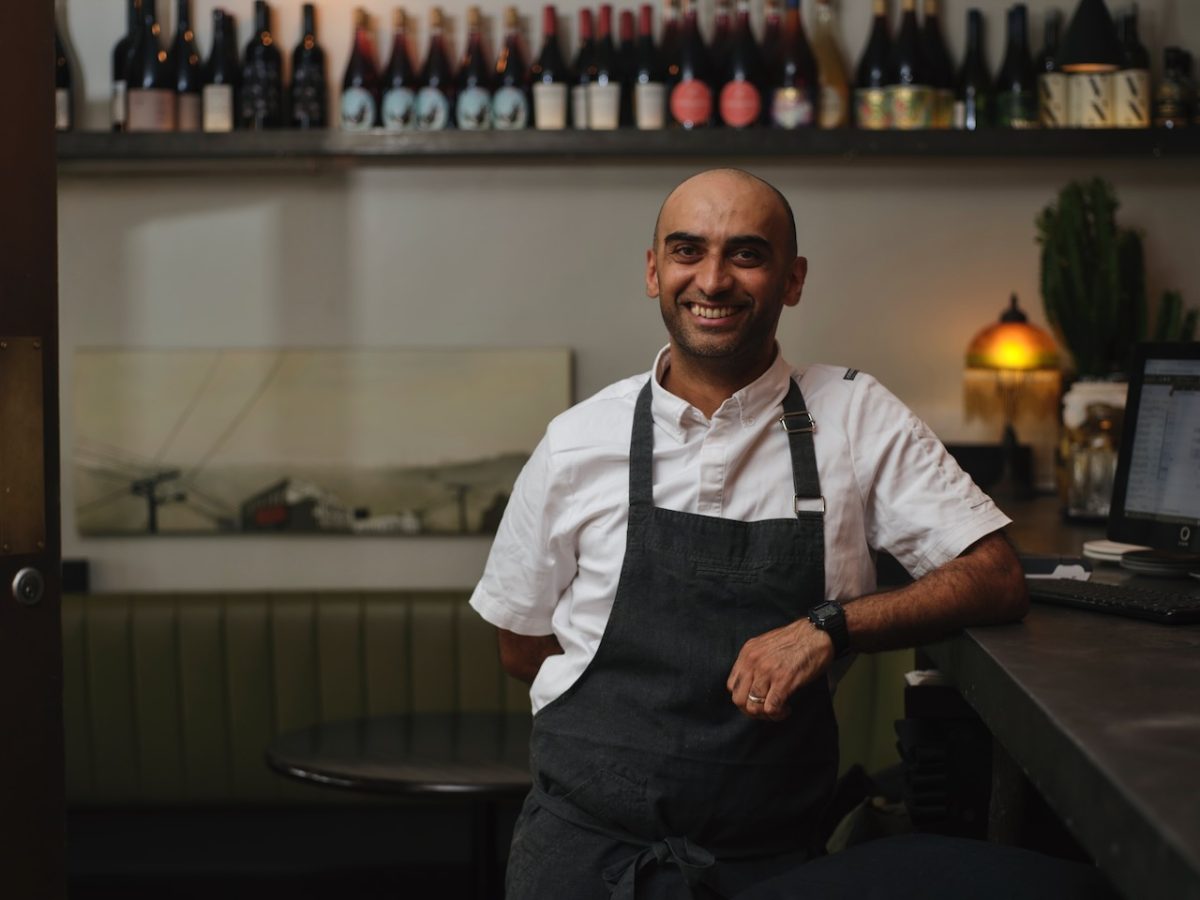A south asian man in a white chefs jacket and dark apron smiles in front of a shelf of wines.