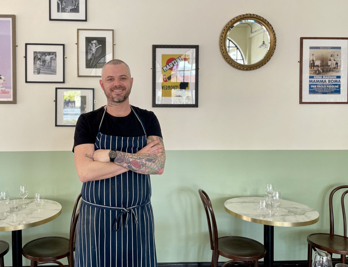 A chef stands with arms crossed in front of a wall decorated with retro posters and a round mirror. 