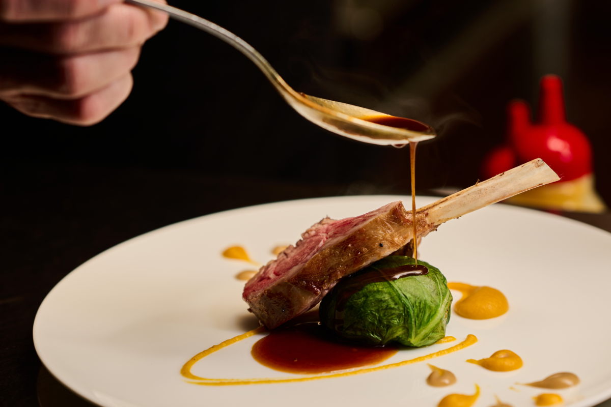 a hand holding a spoon over a dish serving meat and vegetables