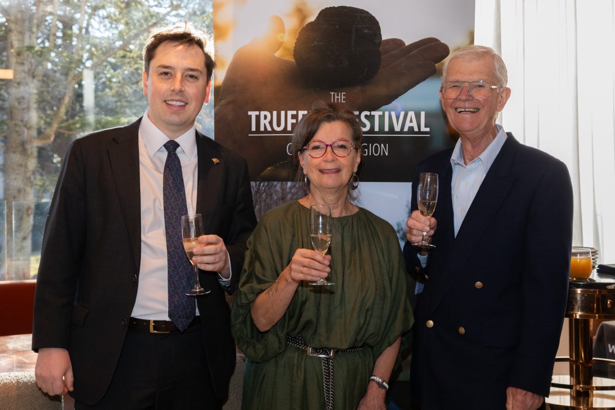 A young man in a suit, a middle aged woman in red glasses and green smock dress, and older man in glasses and a suit hold champagne glasses and smile. They are standing in front of a banner with the image of a truffle.