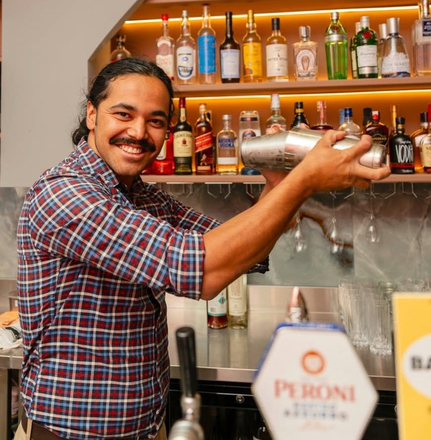 A man with moustache and ponytail shaking a cocktail shaker at a bar.