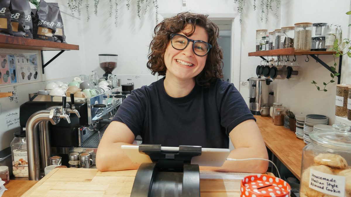 A woman with curly hair, glasses, wearing a black T-shirt smiles behind the counter of a cafe.