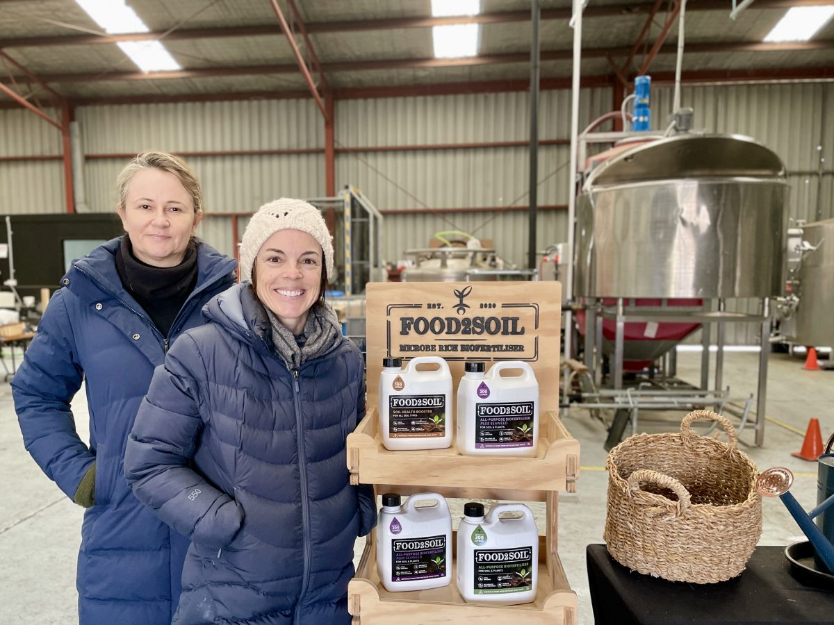 Food2Soil co-CEOs Annabel Schweiger and Josie Grenfell in puffer jackets stand next to a display of bottled biofertiliser.