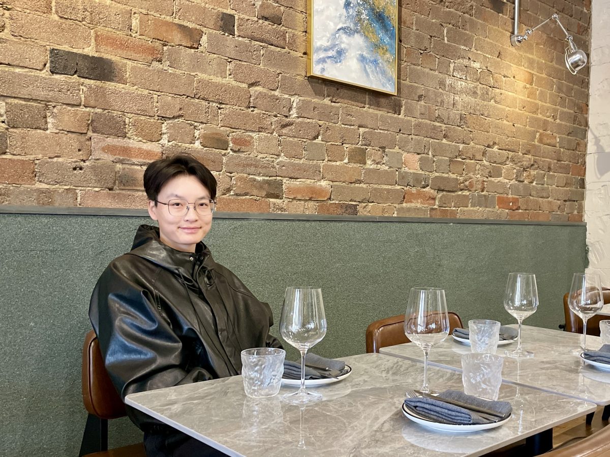 Lina Shou in funky black coat sits at a restaurant table with exposed brick wall behind.