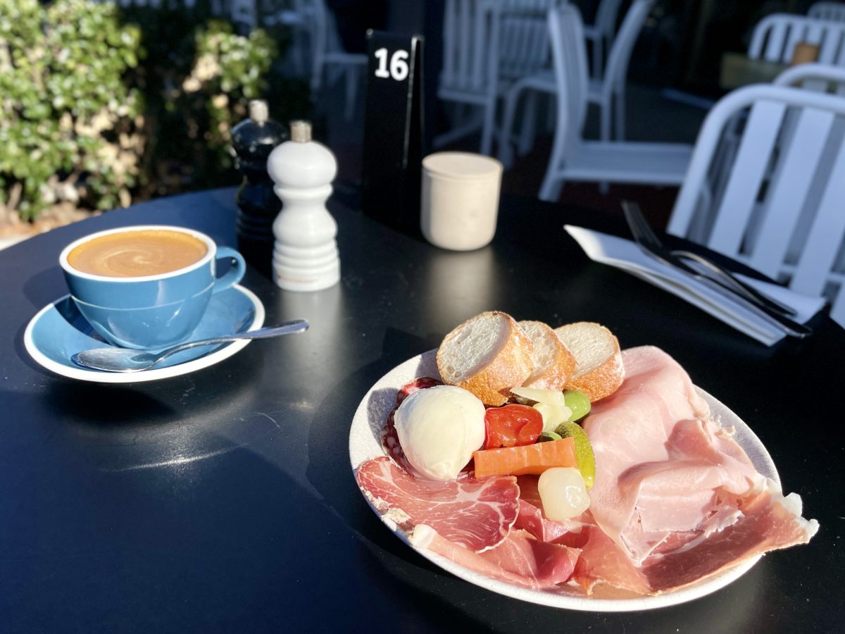 A plate with cured meats, pickles and bread on an outdoor cafe table with a cup of coffee.
