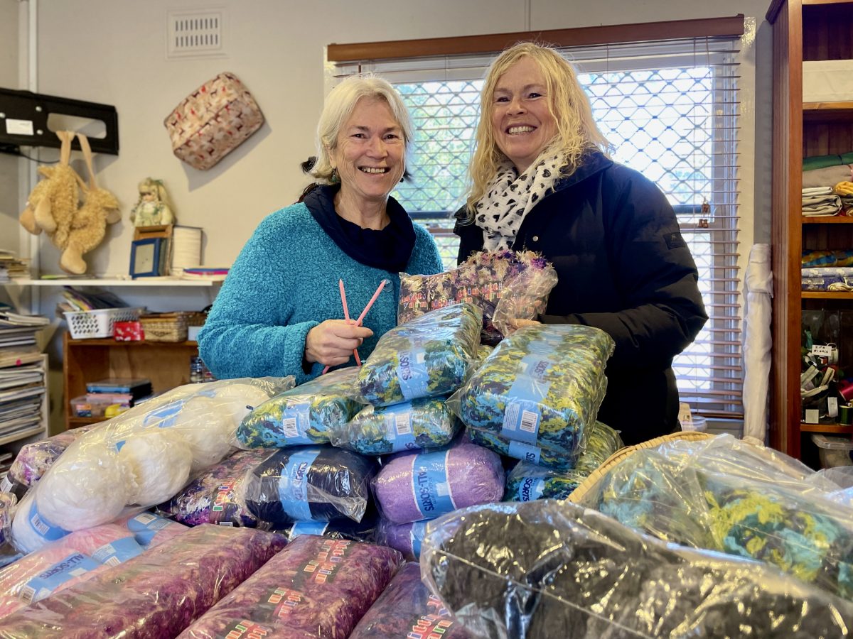 Two woman stand behind a table covered in balls of wool. 