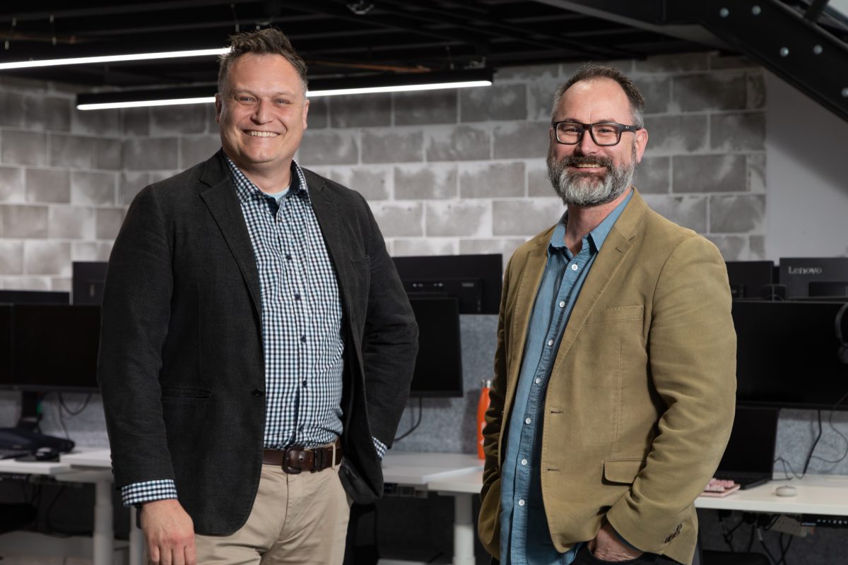 Stephen Oxford and Tony Brine stand in front of computer desks, smiling and looking at the camera