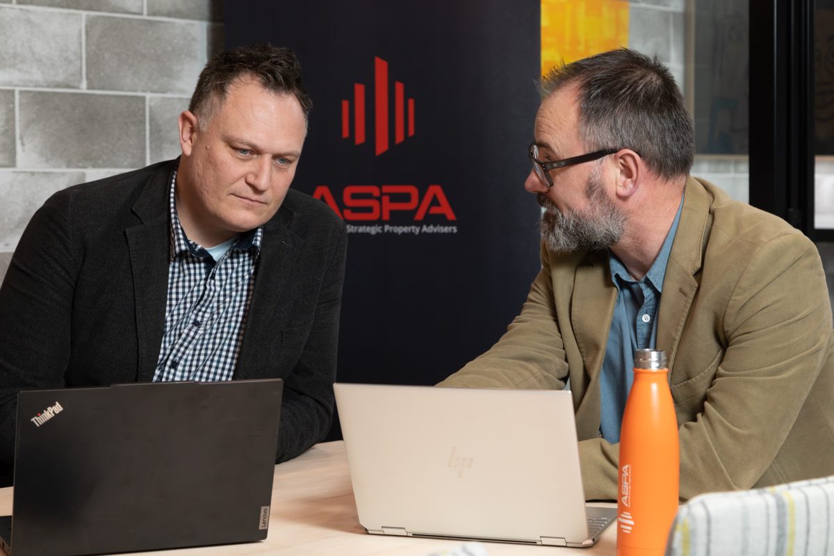Two men seated in front of laptops and talking to each other