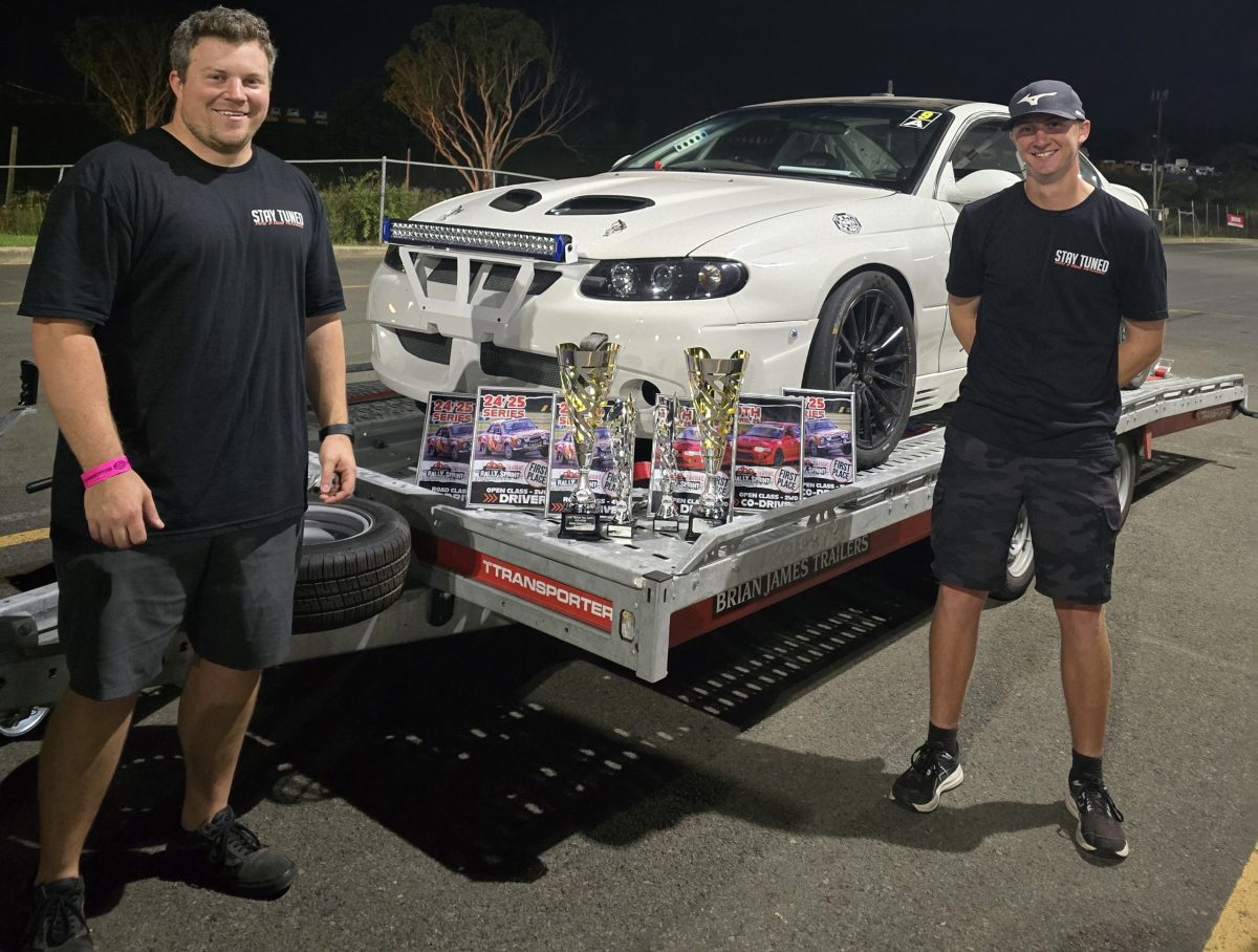 two men standing in front of race car on trailer with trophies