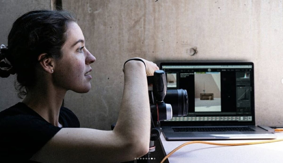 A woman view in profile holds up a camera, in the background a laptop sits on a table. 