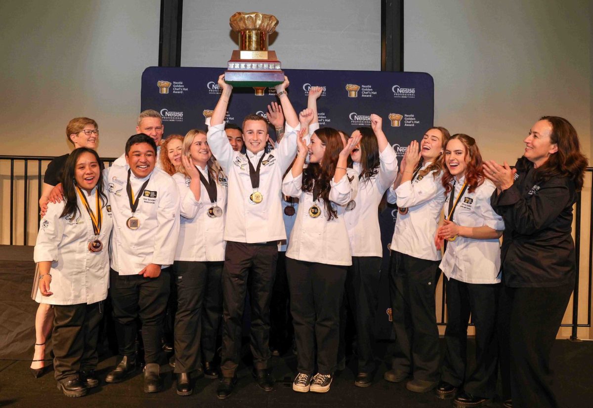 A group photo of young chefs cheering as a young man in the centre hoists a golden chef's hat trophy above his head.