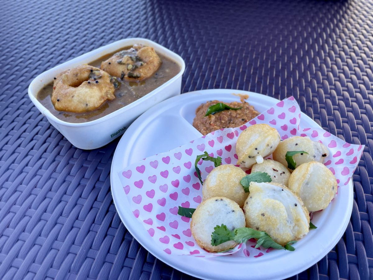 A cardboard plate with small, round rice cakes and a chutney. Behind, a cardboard container with two donut-shaped fritters in a dark red sauce.