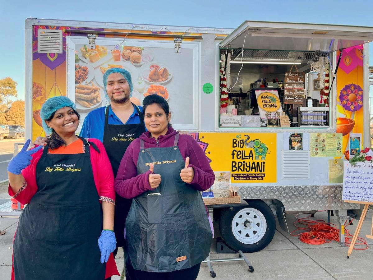 Sisters Nandhini Sri Manogar and Bhuvanee Manogar and Vignesh Ravi stand in front of a colourful food truck which reads "Big Fella Briyani". One woman is giving a double thumbs up and the other is giving a peace sign. All are wearing aprons with Big Fella Briyani branding.