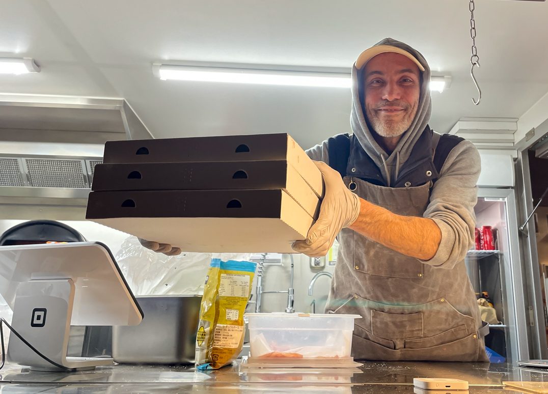 A man in apron, cap and hoodie passes takeaway pizza boxes out of a food truck.