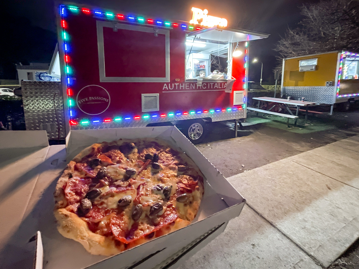 A pizza with salami and olives in a takeaway box, in the background is a pizza truck.