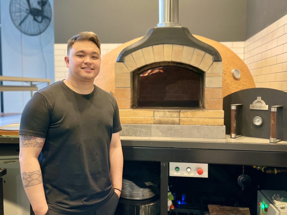 A chef in a black t-shirt stands in front of a pizza oven.