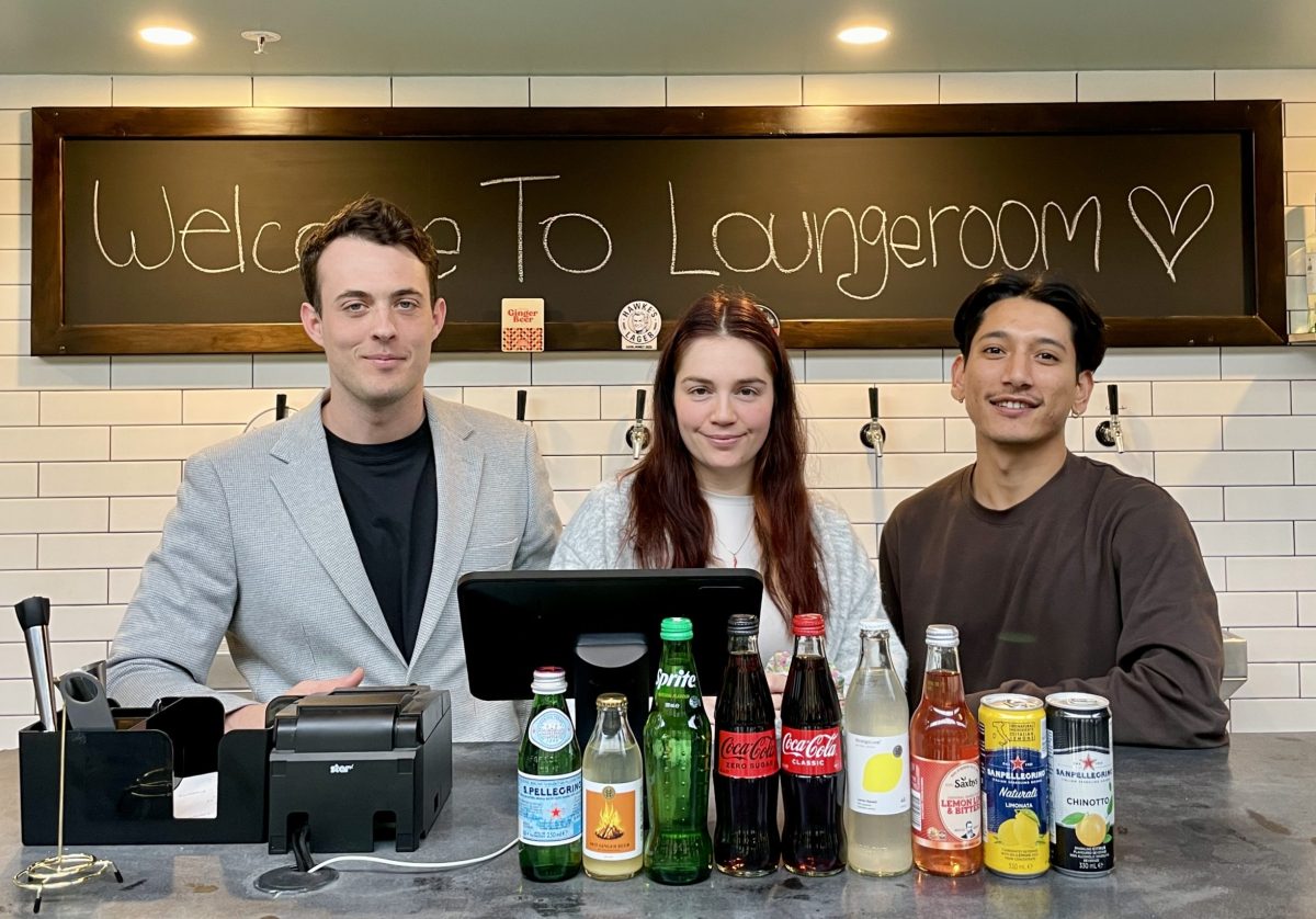 Three people stand in front of a blackboard reading 'Welcome to Lounge Room'