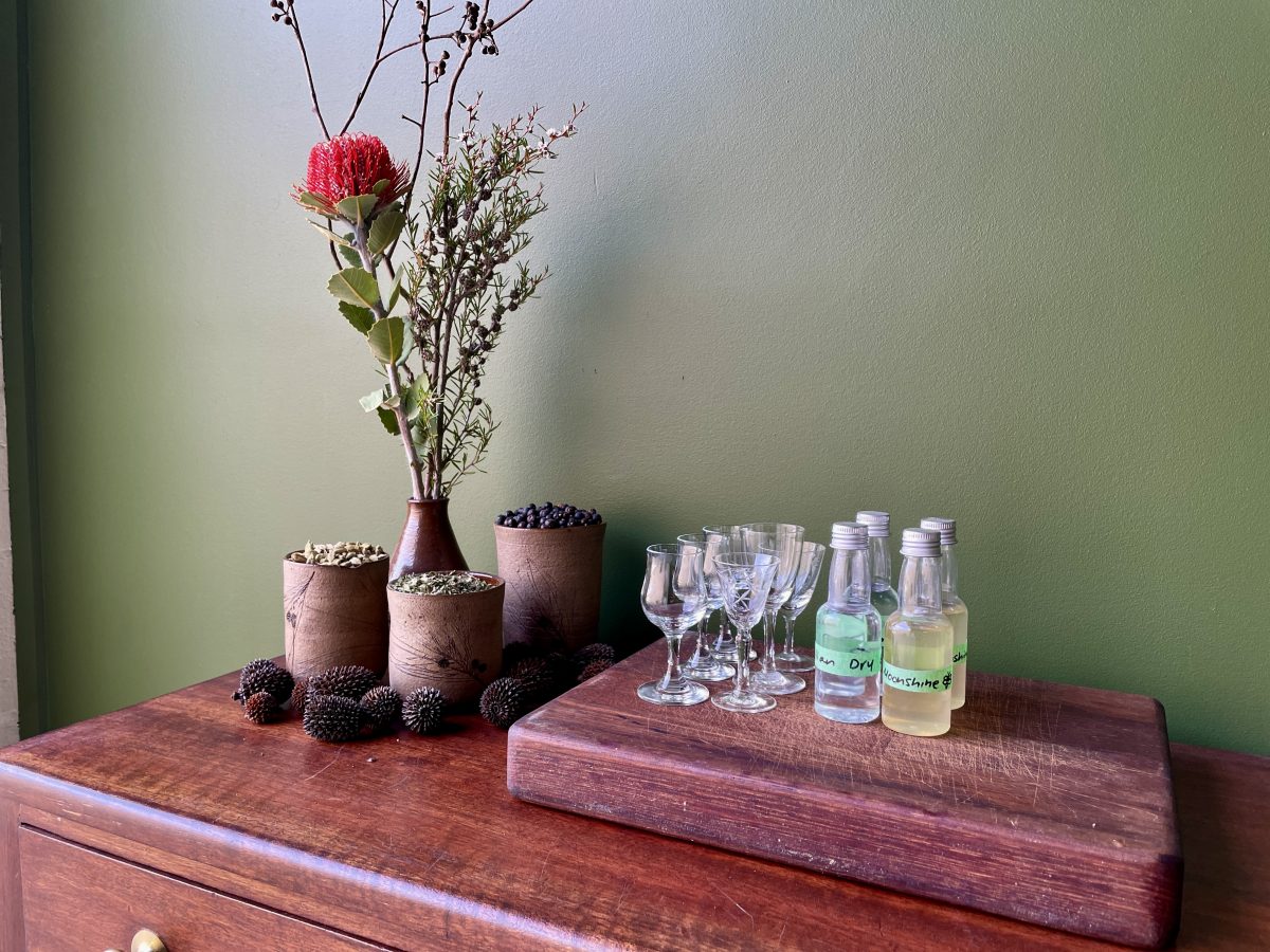 A bunch of native flowers, small bottles of liquor with tape labels and 6 six vintage tasting glasses on a wooden table. 