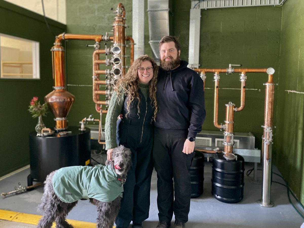 A woman in green jumper and black overalls stands next to a man in black, at their feet a black poodle in green jumper. They are standing in front of a copper still and a green wall.