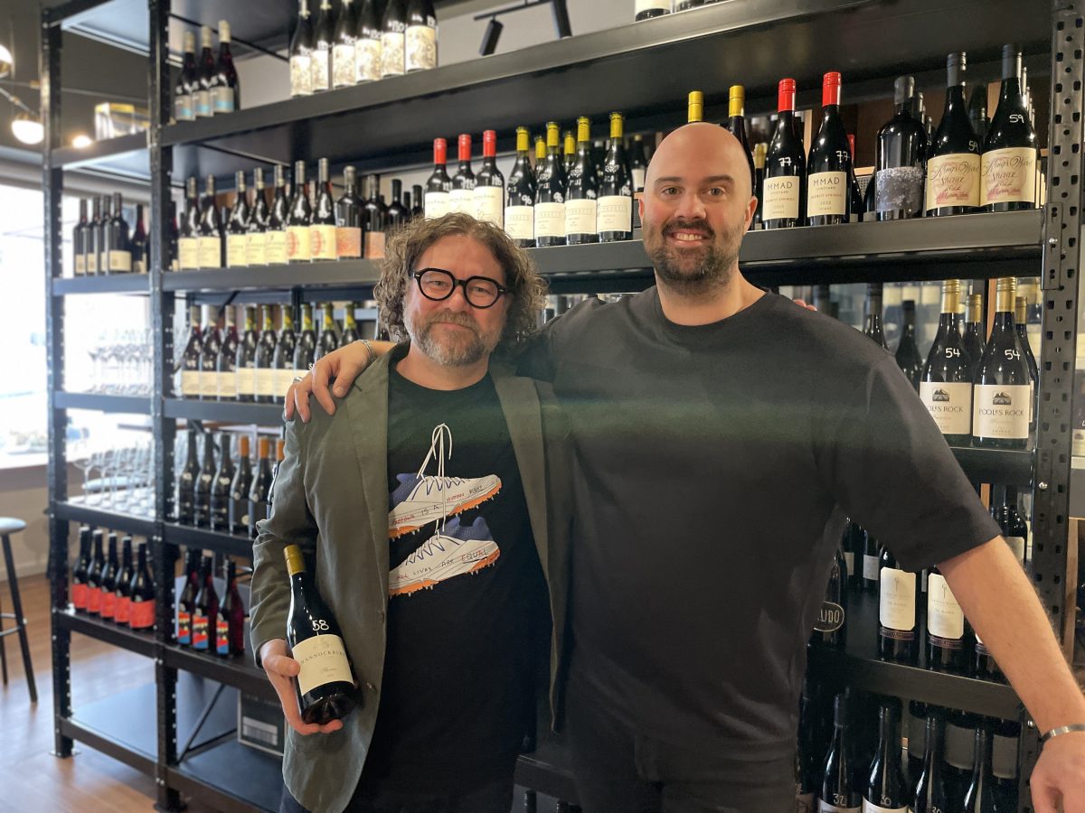 Two men stand in front of a shelf of wine bottles.