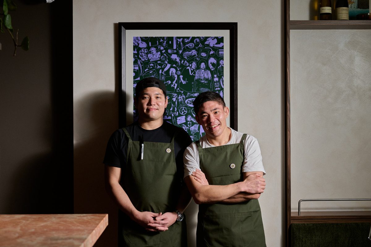 Two brothers in dark green aprons stand in front of a funky green and purple artwork.