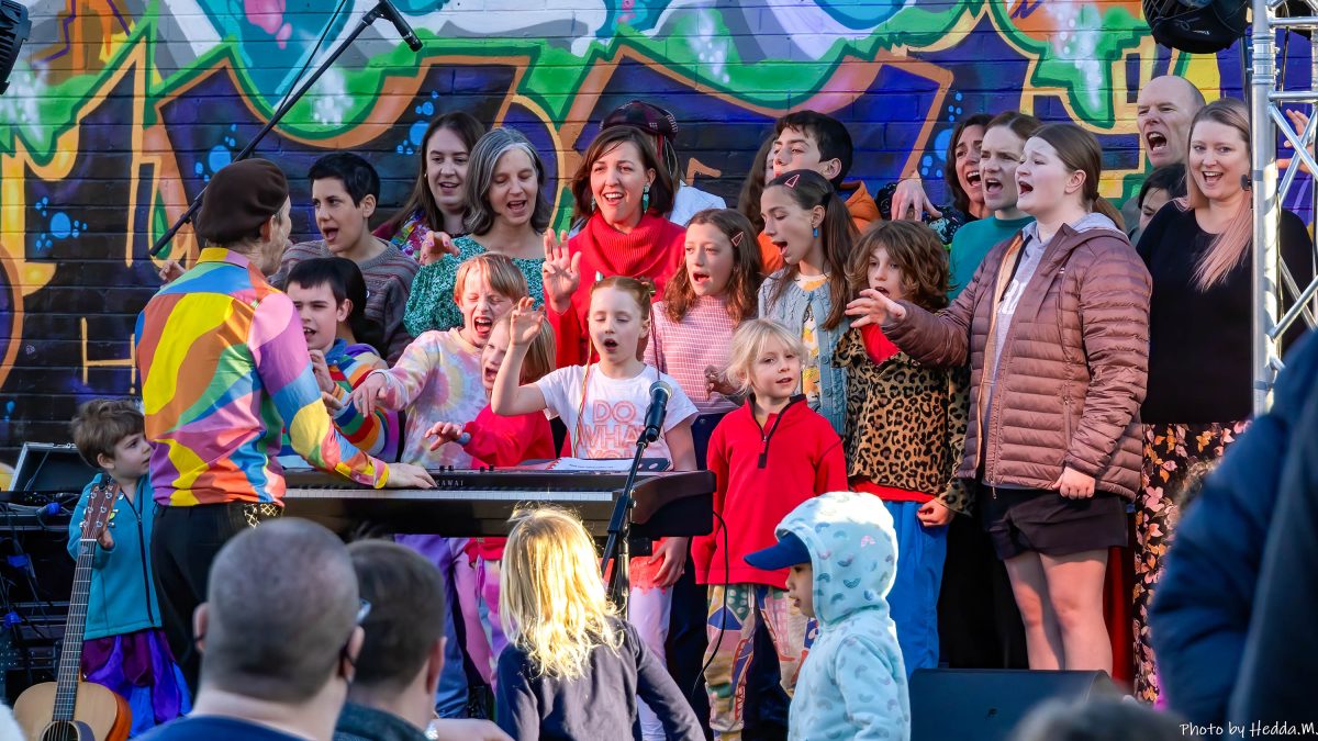 A group of singers of all ages perform in front of a colourful wall.