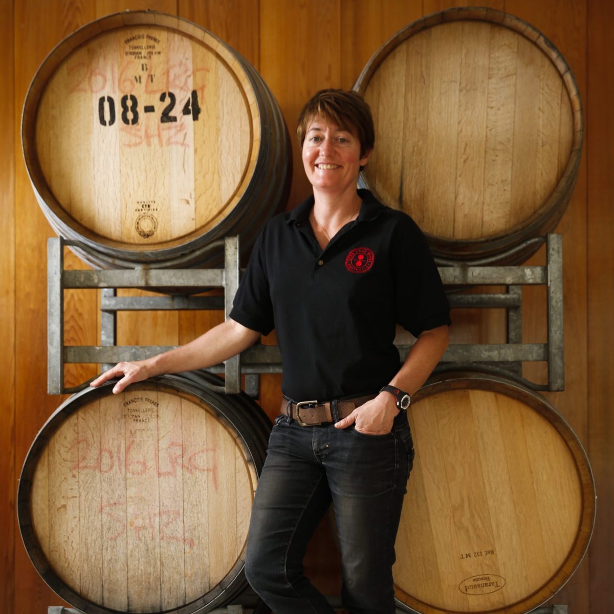 A woman in a black collared shirt stands in front of a stack of wine barrels. 