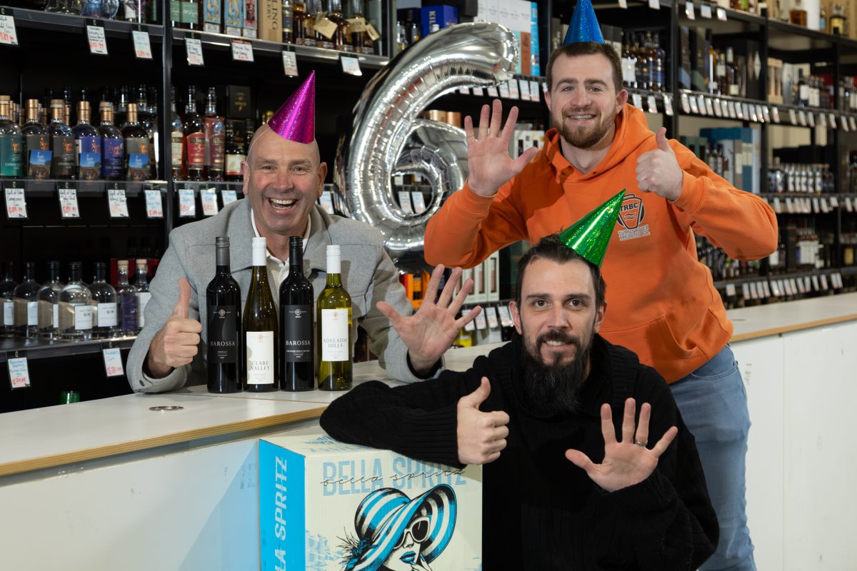 3 people holding up 6 fingers wearing party hats and standing near a counter with wine