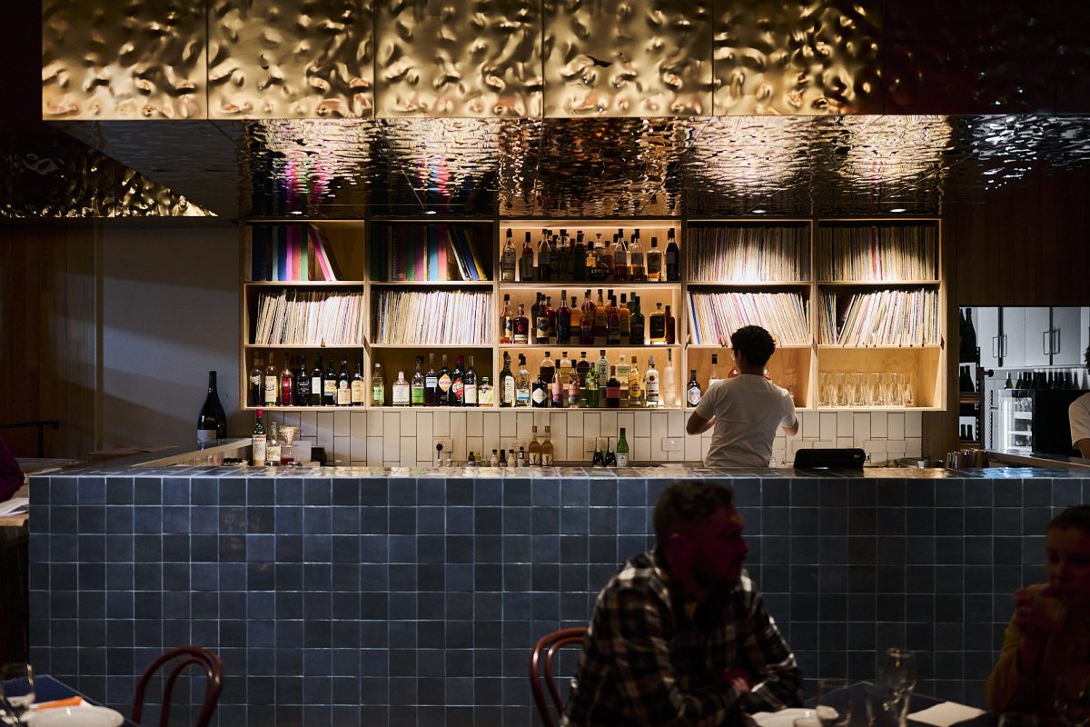 A bartender at a blue-tiled bar with beaten metal roof and shelves of vinyl records.