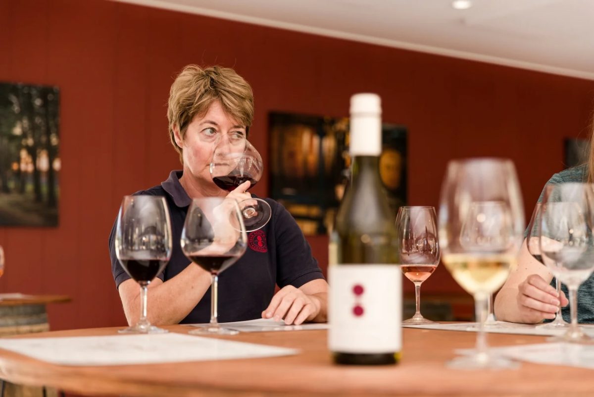 A woman with short hair sniffs a large wine glass. A bottle of Eden Road wine and wineglasses are on the table in the foreground.