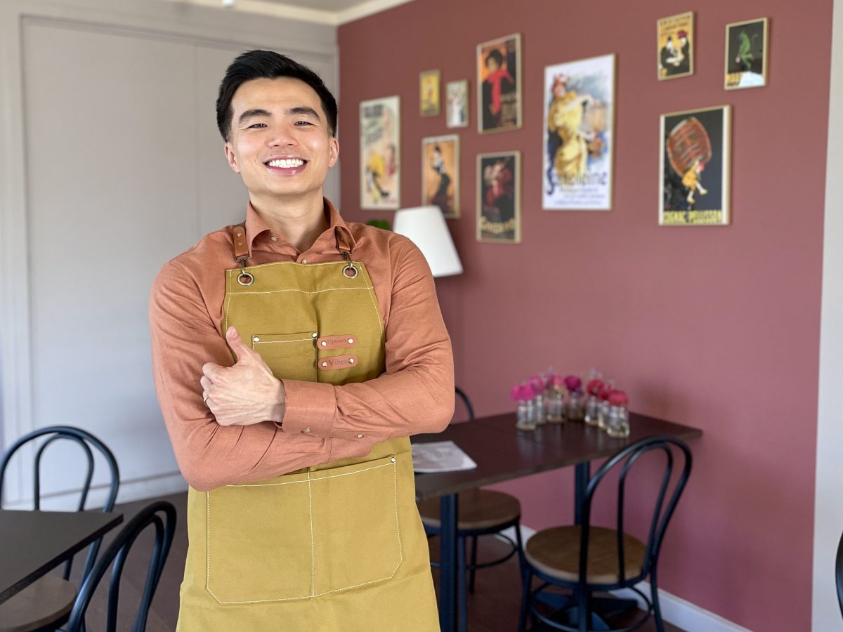 A young Asian man in a peach coloured shirt and yellow Uptown Vibes branded apron smiles and gives a thumbs up to the camera.