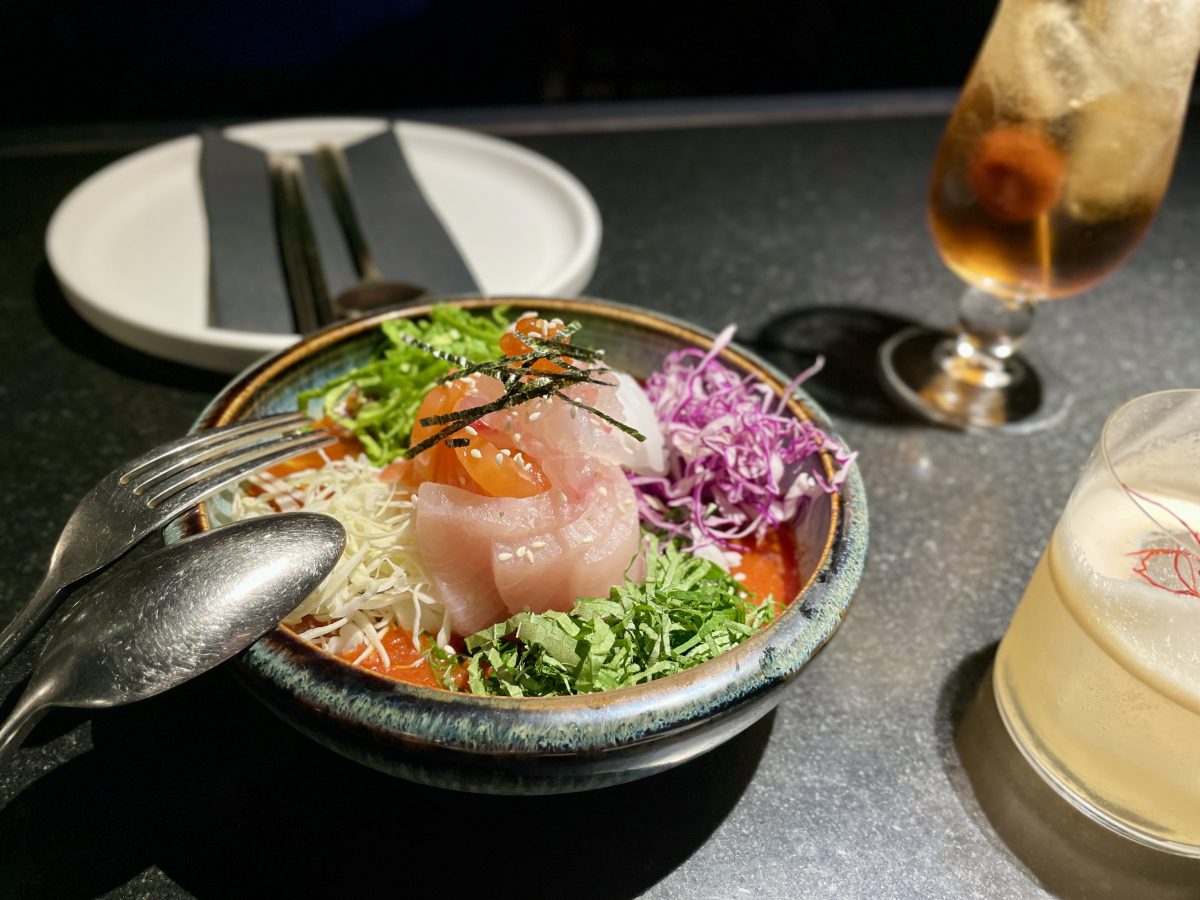 A colourful dish with sashimi fish, and finely sliced vegetables beautifully arranged. On a black table, next to cocktails.