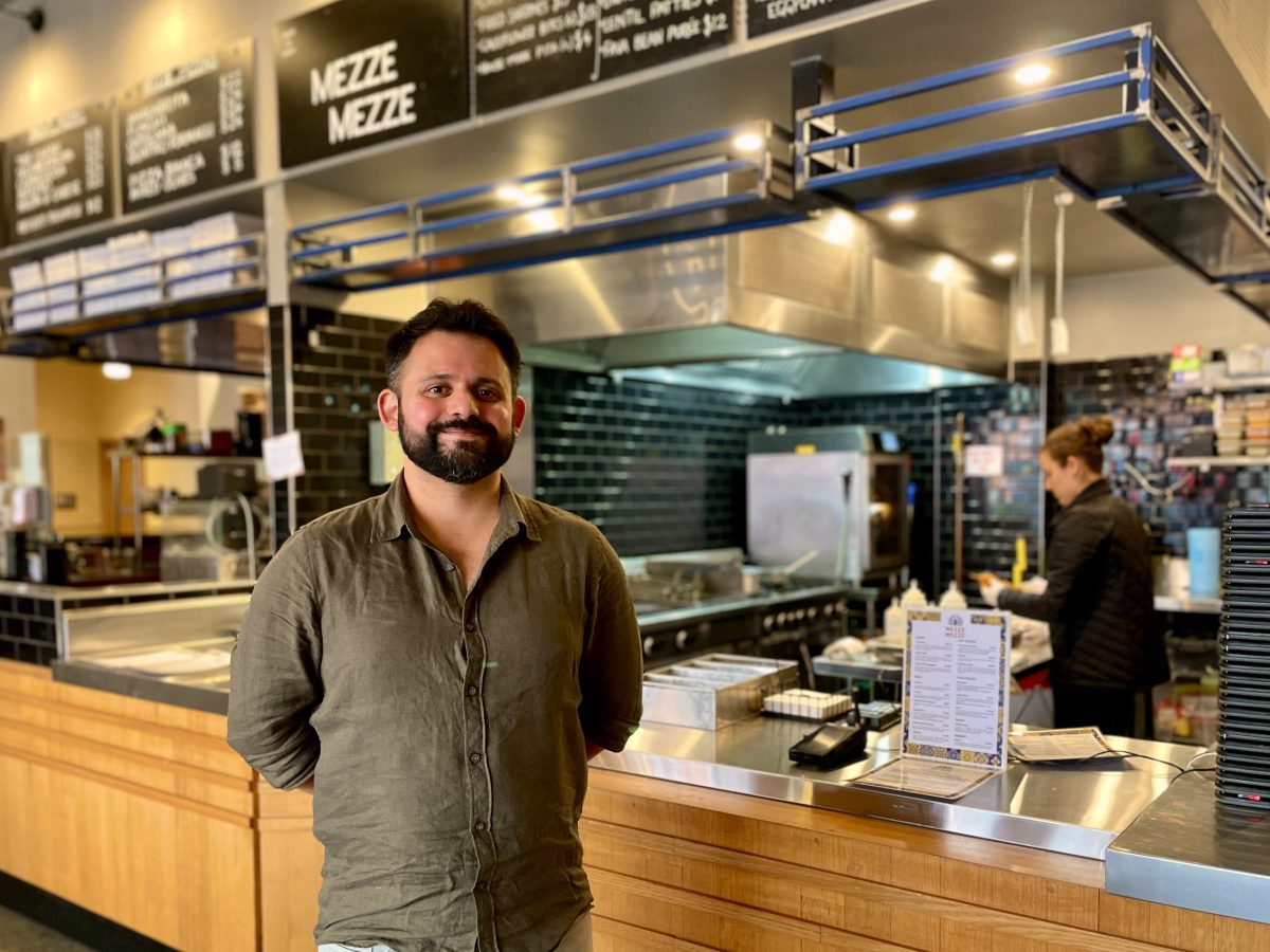 A bearded man in a button down shirt stands in front of an open kitchen with a sign reading Mezze Mezze.
