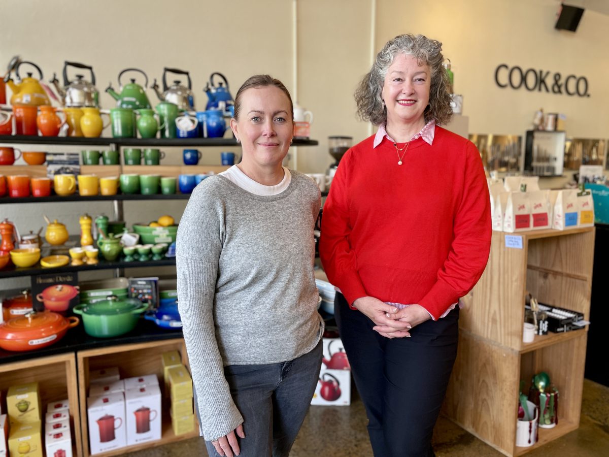Cook and Co owners Kate Wellington and Claire Smith stand in front of a shelf of colourful kitchenware. In the background, the words Cook & Co are on the wall in black lettering.