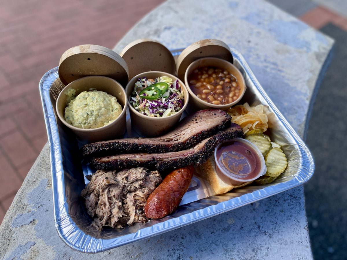 A large aluminium tray of tasty looking barbecued meats, with round containers of side dishes.