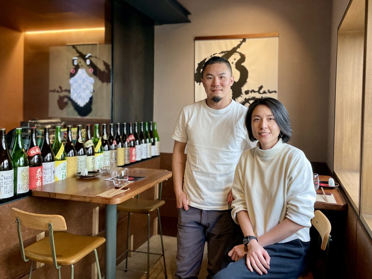 A man with goatee and ponytail stands in white shirt, woman in white sweater sits on a stool beside him. In the background, bottles of sake, a calligraphy image on the wall and neon logo of ramen daddy.